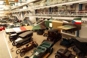 Ein großer Museumsraum mit verschiedenen Oldtimern, Flugzeugen, Fahrrädern und Informationsschildern, bei denen Besucher die Ausstellungsstücke betrachten.