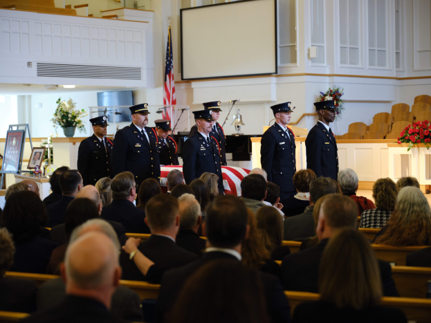 Eine Gruppe uniformierter Personen mit Mützen, die vor sitzenden Trauernden bei einer Beerdigungszeremonie stehen, mit Blumensträußen, einer Fahne, einem Bildschirm und einer Wand im Hintergrund.