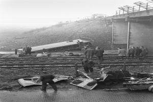Eine Schwarz-Weiß-Szene eines Zugunglücks auf den Gleisen mit einer Gruppe von Menschen um das Wrack herum, einem Fahrzeug im Vordergrund und einer Brücke, Polen, Drähte und Himmel im Hintergrund.