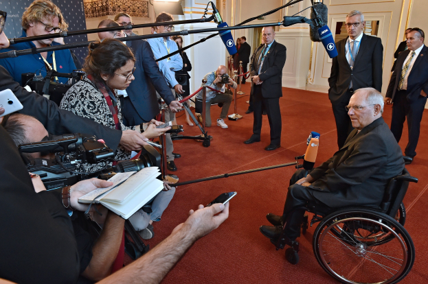 Ein Mann im Rollstuhl spricht zu Reportern mit Kameras, Mikrofonen und Handys, mit einem Banner, Scheinwerfern und einer Wand im Hintergrund.