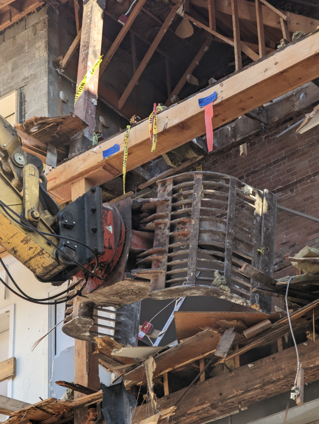 Ein Bagger arbeitet neben einem teilweise abgerissenen Gebäude, das von Holzplanken und Schutt umgeben ist.