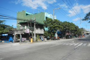 Eine Stadtstraße mit einem grünen Gebäude, fahrende Fahrzeuge, Fußgänger auf dem Gehweg und Strommasten mit Drähten vor einem bewölkten Himmel.