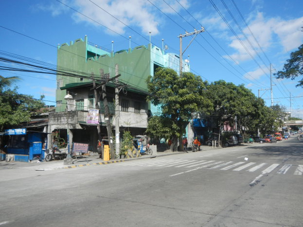 Eine Stadtstraße mit einem grünen Gebäude, fahrende Fahrzeuge, Fußgänger auf dem Gehweg und Strommasten mit Drähten vor einem bewölkten Himmel.