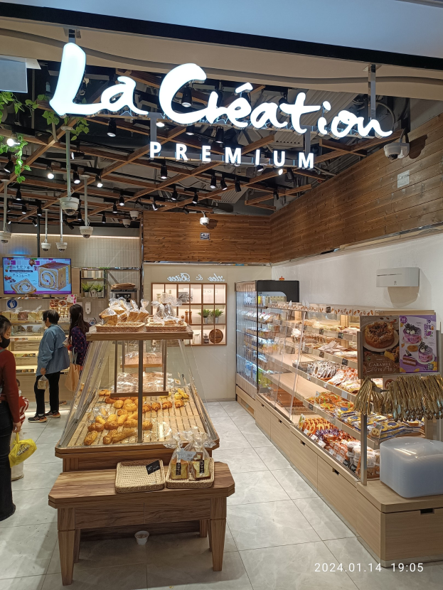 Eine Gruppe von Menschen in einem Café mit einer Auslage voller Lebensmittel, einem Tisch mit Körben, einem Schild, einem Fernseher, Pflanzen, Deckenleuchten und einem Dach.