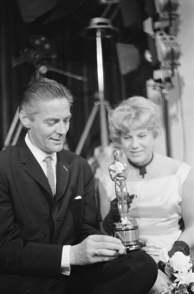 Ein Mann in einem schwarzen Blazer, weißem Hemd und Krawatte hält eine Oscar-Statue, während er neben einer Frau in Abendgarderobe sitzt, die einen Blumenstrauß hält, beide in Schwarz-Weiß.