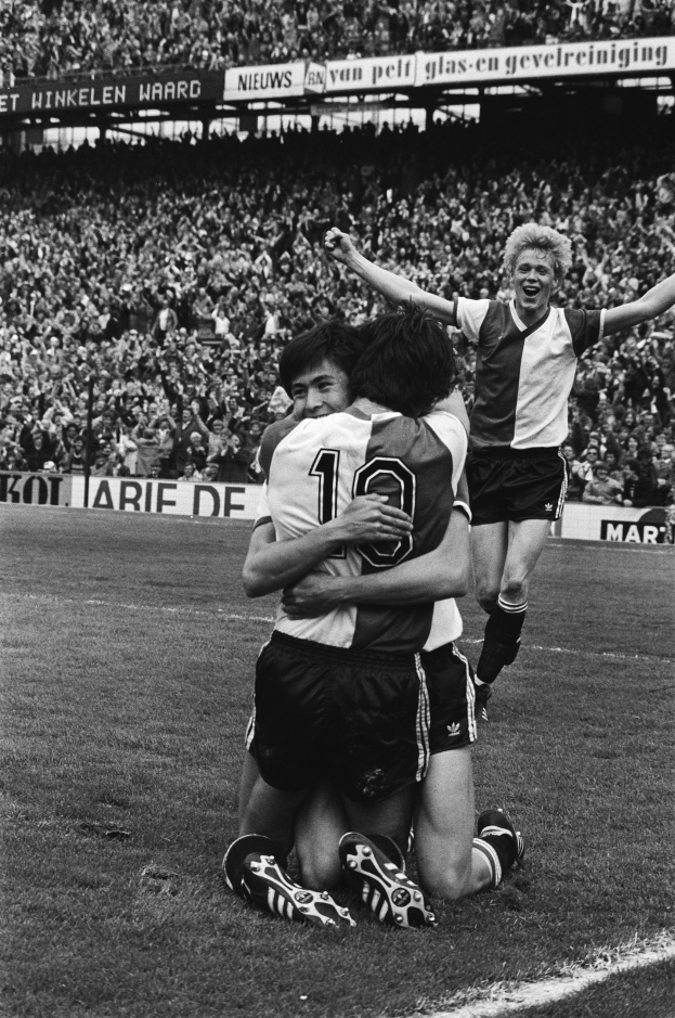 Drei Fußballspieler feiern Tor auf dem Boden, mit jubelnder Menge und schwenkenden Fahnen im Hintergrund, in einem schwarz-weißen Stadion Setting.