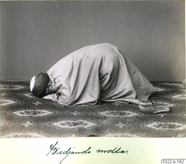 Ein altes Schwarz-Weiß-Foto eines Mannes im Gebet, der ein Tuch und eine Kappe trägt, mit der Aufschrift "Bredjande Molla, die erste muslimische Frau der Welt" unten.
