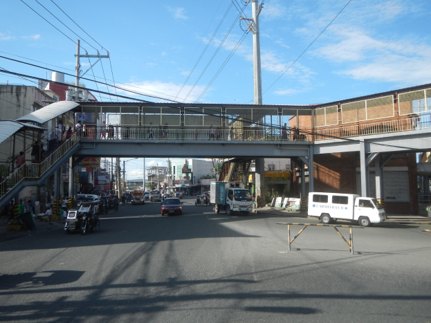Stadtstraße mit Fahrzeugen, eine Fußgängerbrücke mit Menschen, Strommasten mit Kabeln, Gebäude und ein bewölkter Himmel.