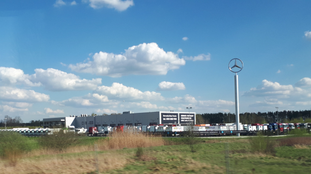 Mercedes-Benz-Fabrik in Deutschland mit Fahrzeugen auf der Strasse, Gruen, Gebaeuden und Logo-Pfahl unter einem bewolkten Himmel.