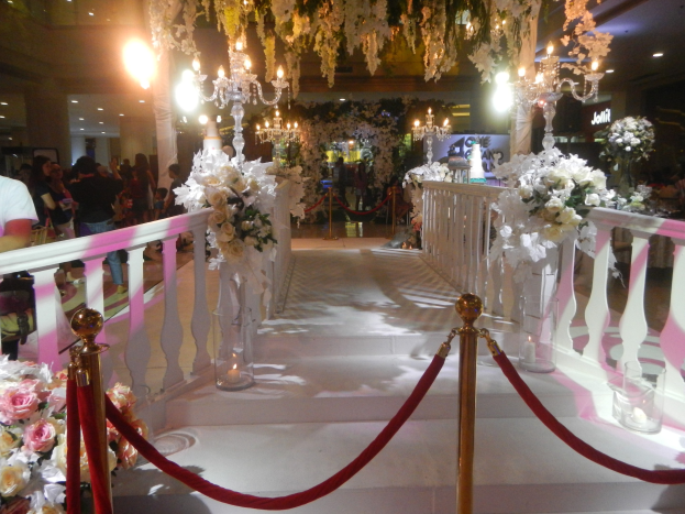 Eine Hochzeitszeremonie in einem Einkaufszentrum mit weißem Teppich, dekorativen Geländern, Blumenschmuck, Kronleuchtern und Deckenbeleuchtung, bei der auch einige Taschen tragenden Personen anwesend sind.