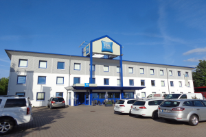 Ein modernes Gebäude mit einer Tafel, auf der "ibis budget frankfurt airport" steht, geparkte Autos, Topfpflanzen, eine Straßenlaterne, Bäume und ein bewölkter Himmel.