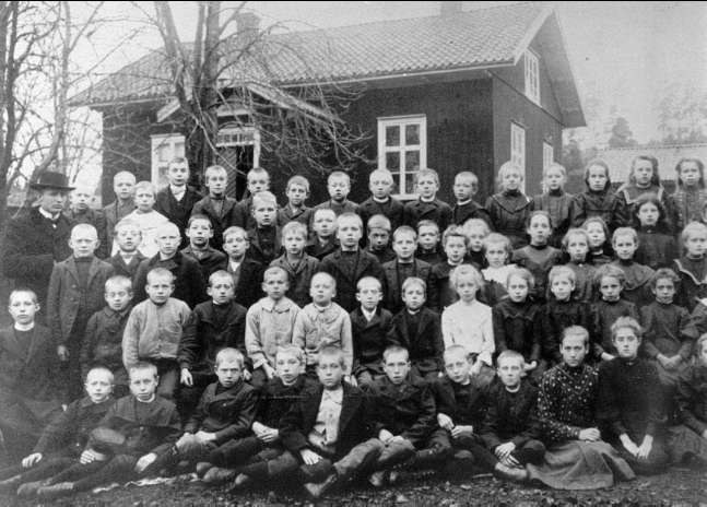 Schwarz-weiß-Foto einer Gruppe von Kindern, die vor einem Haus mit Fenstern posieren, umgeben von Bäumen und einem klaren Himmel, mit dem Text "1930er Schulkinder" unten.