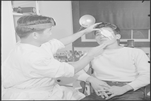 Ein Schwarz-Weiß-Foto eines Mannes, der in einem Stuhl sitzt, während ein Arzt neben ihm seine Augen mit einem tragbaren Objekt untersucht, mit einem Tisch und einem Fenster mit einem Vorhang im Hintergrund.
