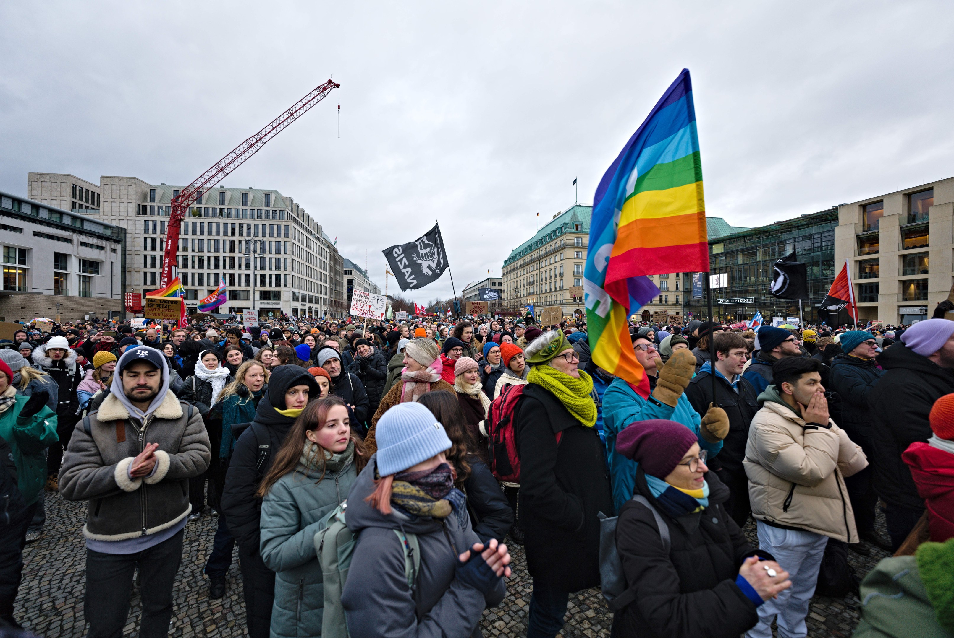 Eine große Gruppe von Menschen steht vor einem Gebäude und hält Fahnen und Schilder mit der Aufschrift 'Lgbtq+ rights march in Berlin', einige tragen Mützen und Taschen, im Hintergrund sind Gebäude mit Fenstern, ein Kran und ein bewölkter Himmel zu sehen.