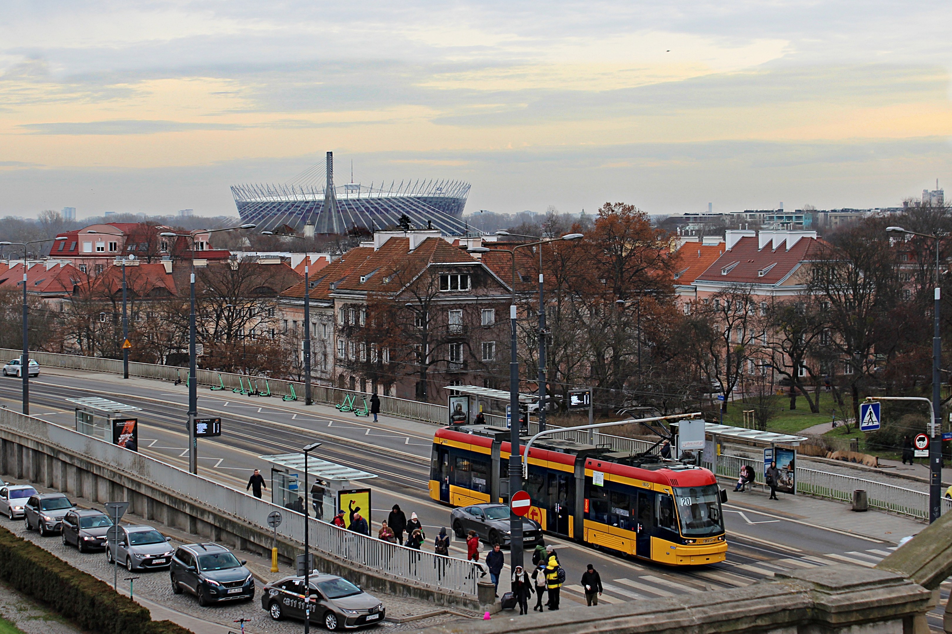 Eine gelbe und schwarze Straßenbahn fährt auf einer von hohen Gebäuden gesäumten Stadtstraße, umgeben von Autos, Fußgängern, Laternen, Schildern, Bäumen und Gras, mit bewölktem Himmel im Hintergrund.