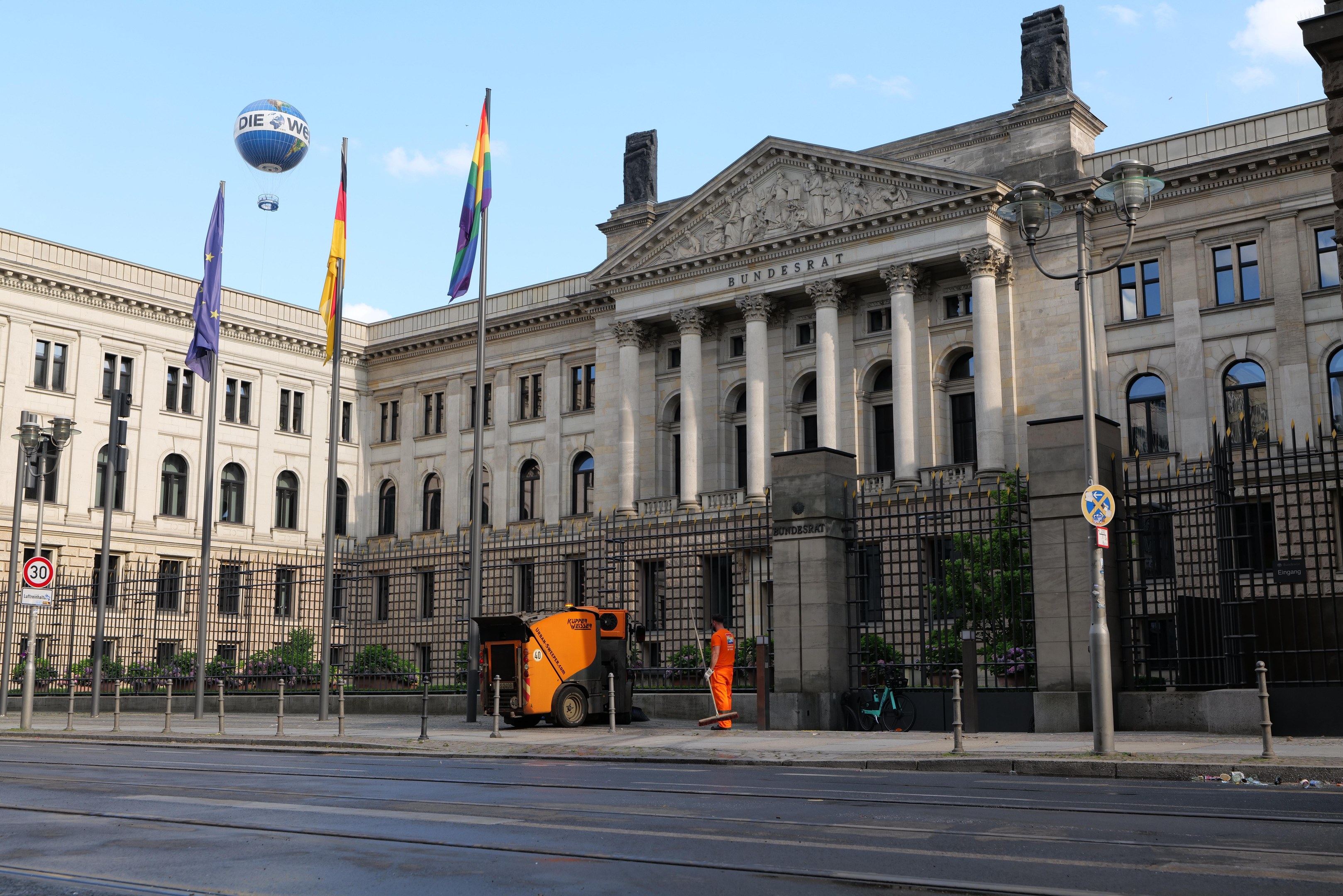 Großes Gebäude mit zahlreichen Fenstern, als Bundestag in Berlin, Deutschland identifiziert, umgeben von Straßeninfrastruktur und Fußgängern, mit Flaggen und Bäumen im Vordergrund und einem bewölkten Himmel im Hintergrund.