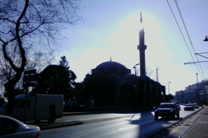 Eine Moschee steht in der Mitte einer Stadtstraße, umgeben von Gebäuden, Straßenlaternen, Strommasten, Stromkabeln, Kraftfahrzeugen und Bäumen unter einem klaren blauen Himmel.