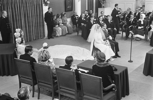 Schwarz-weiß-Foto einer Hochzeitszeremonie mit sitzenden und stehenden Menschen, einem Mann mit Mikrofon, einer Frau mit Blumenstrauß, einem Podium mit Mikrofon, Blumenbouquets, einem Vorhang, einem Foto Rahmen an der Wand und einer Tür im Hintergrund.
