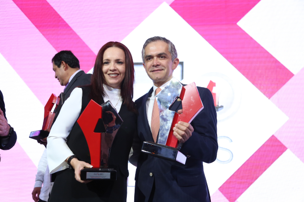 Ein Mann und eine Frau stehen nebeneinander auf einer Bühne, jeder hält eine Trophäe und lächelt, mit einem Bildschirm im Hintergrund.