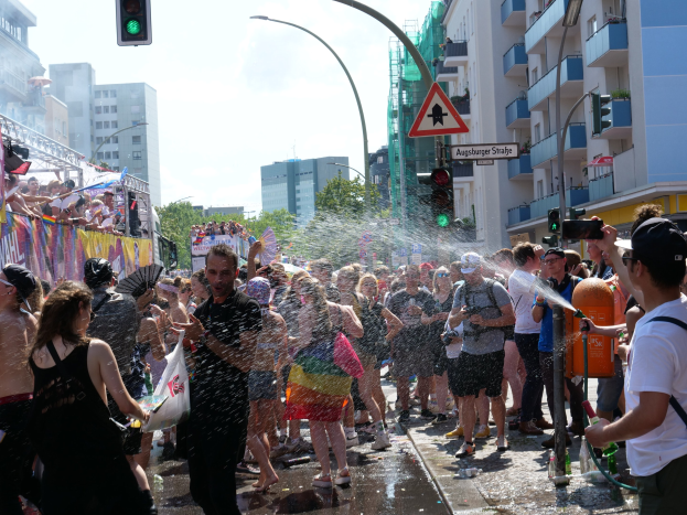 Menschen auf einer Christopher Street Parade, die sich gegenseitig mit Wasser bespritzen und Gegenstände halten, mit einem Banner links und Gebäuden, Bäumen und Straßenelementen im Hintergrund.