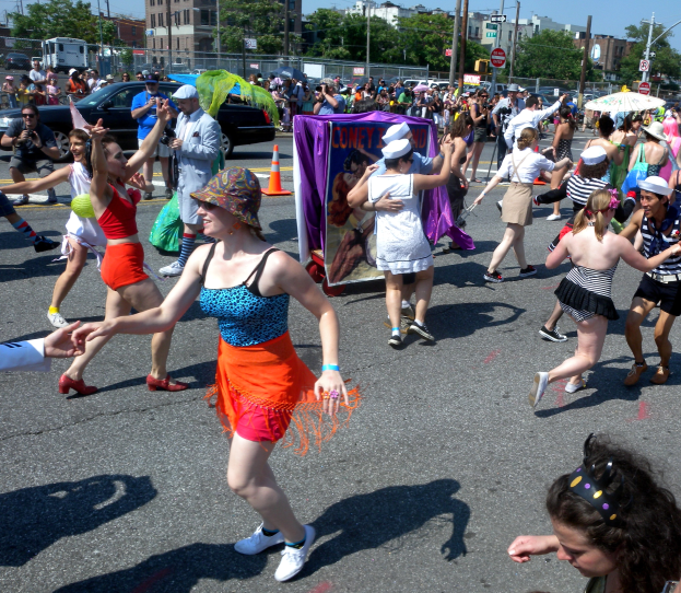 Eine Gruppe von Menschen, die auf der Straße in einer Parade marschieren, einige mit Mützen und Schirmen, andere tanzend, mit einem Banner, Verkehrskegeln, Fahrzeugen, Laternenmasten, Schildern, Bäumen, Gebäuden und einem klaren blauen Himmel im Hintergrund.