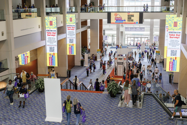 Eine große Gruppe von Menschen, die durch ein großes Gebäude mit Bannern, Topfpflanzen, Rolltreppen, Geländern, Säulen, Texttafeln, Glastüren und Glastüren geht, einige tragen Taschen, wahrscheinlich an der Nationalen Buchmesse teilnehmend.