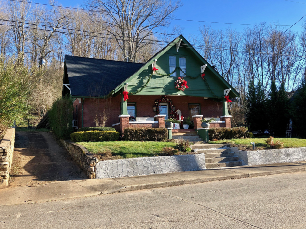 Ein gr├╝nes Haus mit einem Kranz an der Vorderseite, umgeben von Pflanzen, Gras, Treppen, einer Wand, einer Baumgruppe, einigen Dr├Ąhten und einem bew├Âlktem Himmel, das sich im Herzen der Stadt befindet und zur Miete verf├╝gbar ist.