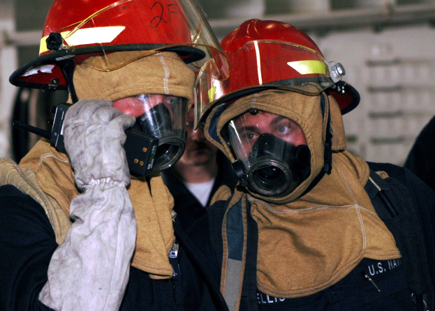 Zwei Feuerwehrleute in Gasmasken und Helmen, einer hält ein Funkgerät, stehen neben einer Wand mit Rohren im Hintergrund.