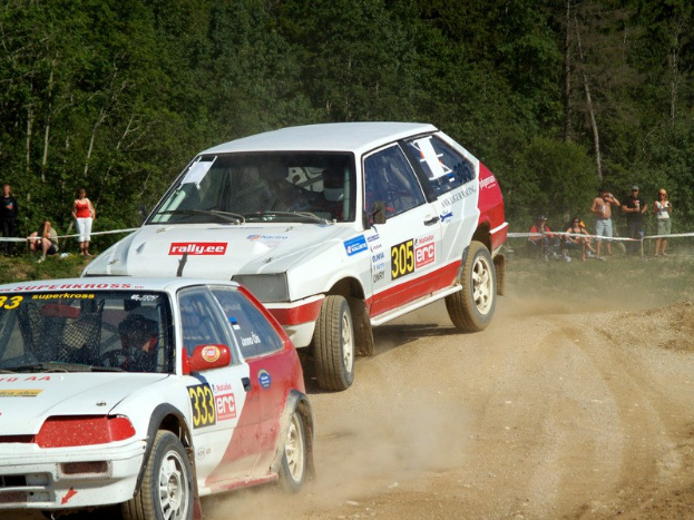 Zwei Autos mit Text und Zahlen darauf fahren auf einer Schotterstraße an einer Menschenmenge vorbei, einige sitzen und einige stehen, mit einem Geländer und Bäumen im Hintergrund.