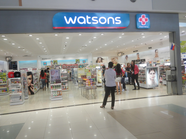Eine Watsons Apotheke in einem Einkaufszentrum mit Menschen, Regalen, Schildern, einer Wanduhr und Deckenleuchten.