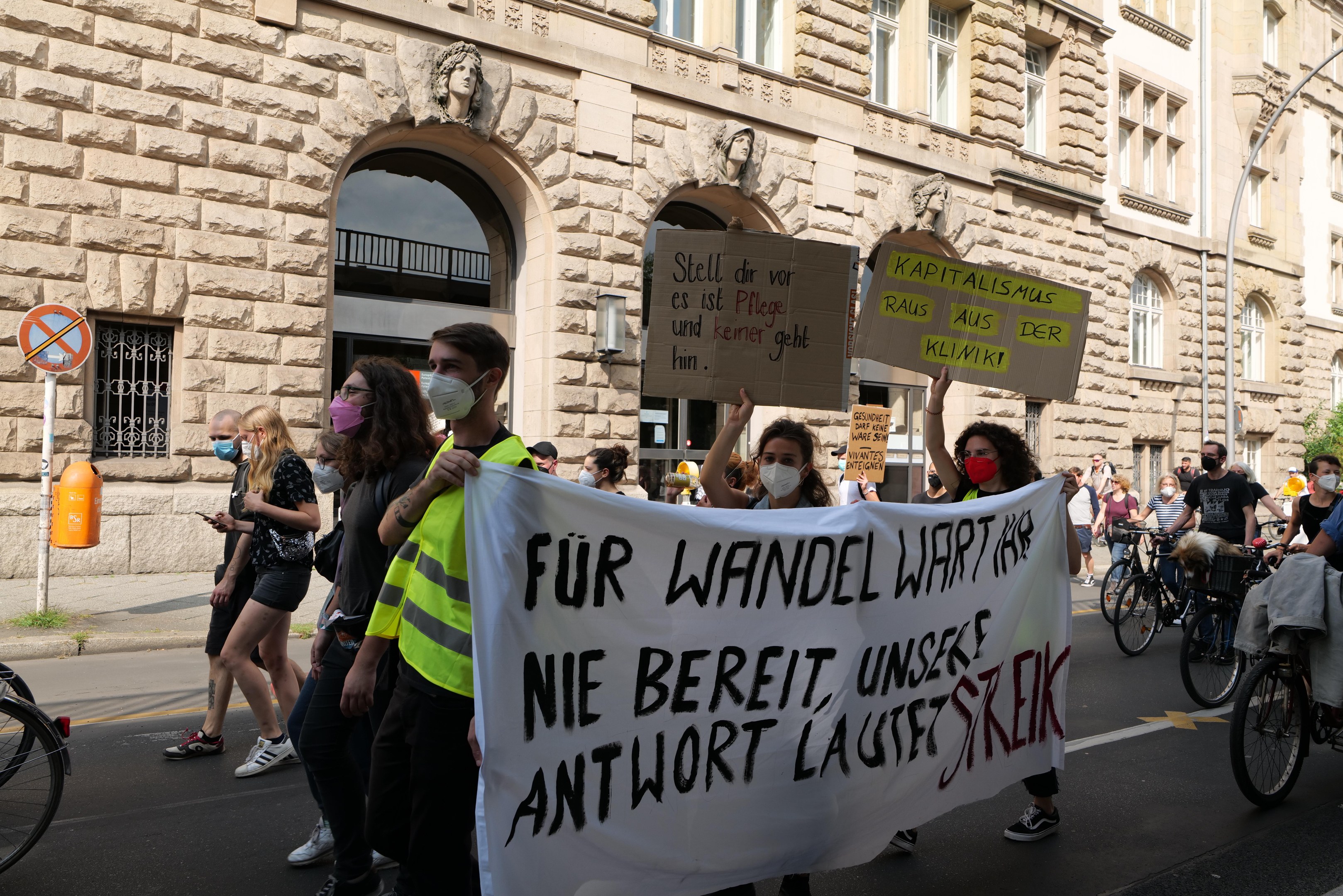 Eine Gruppe von Menschen, die auf einer Straße marschieren, einige mit Schildern und andere mit Fahrr√°dern, mit einem Gebäude im Hintergrund, das bogige Fenster, Pfeiler und Skulpturen aufweist.