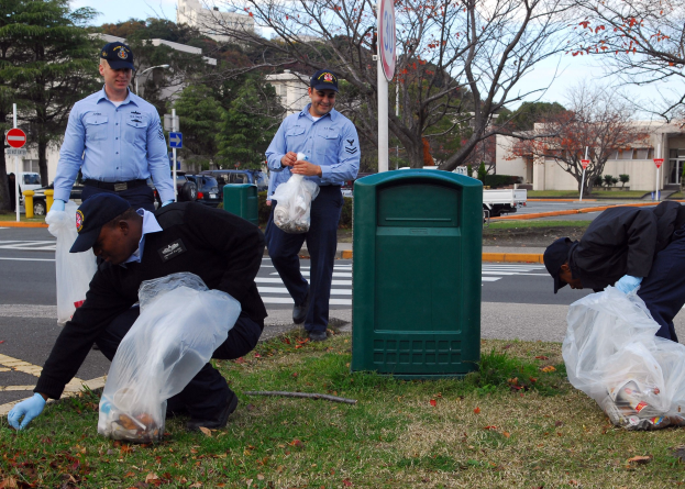 Drei Polizisten in Uniform sammeln Müll am Straßenrand mit Bäumen, Gebäuden und Fahrzeugen im Hintergrund.