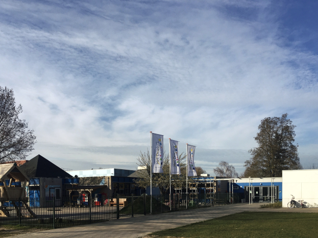 Großes Gebäude mit Metallzaun, Rasenfläche, Weg, Bäumen, Pfählen, Bannern, einem Fahrrad, nahen Häusern und einem bewölkten Himmel, als neuer Spielplatz an der Schule identifiziert.